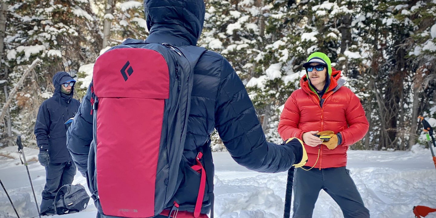 Two people in ski gear and in the snow practice using their beacon
