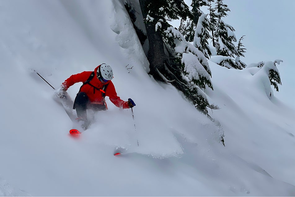 A man in a red jacket skiing down a steep mountain in powder