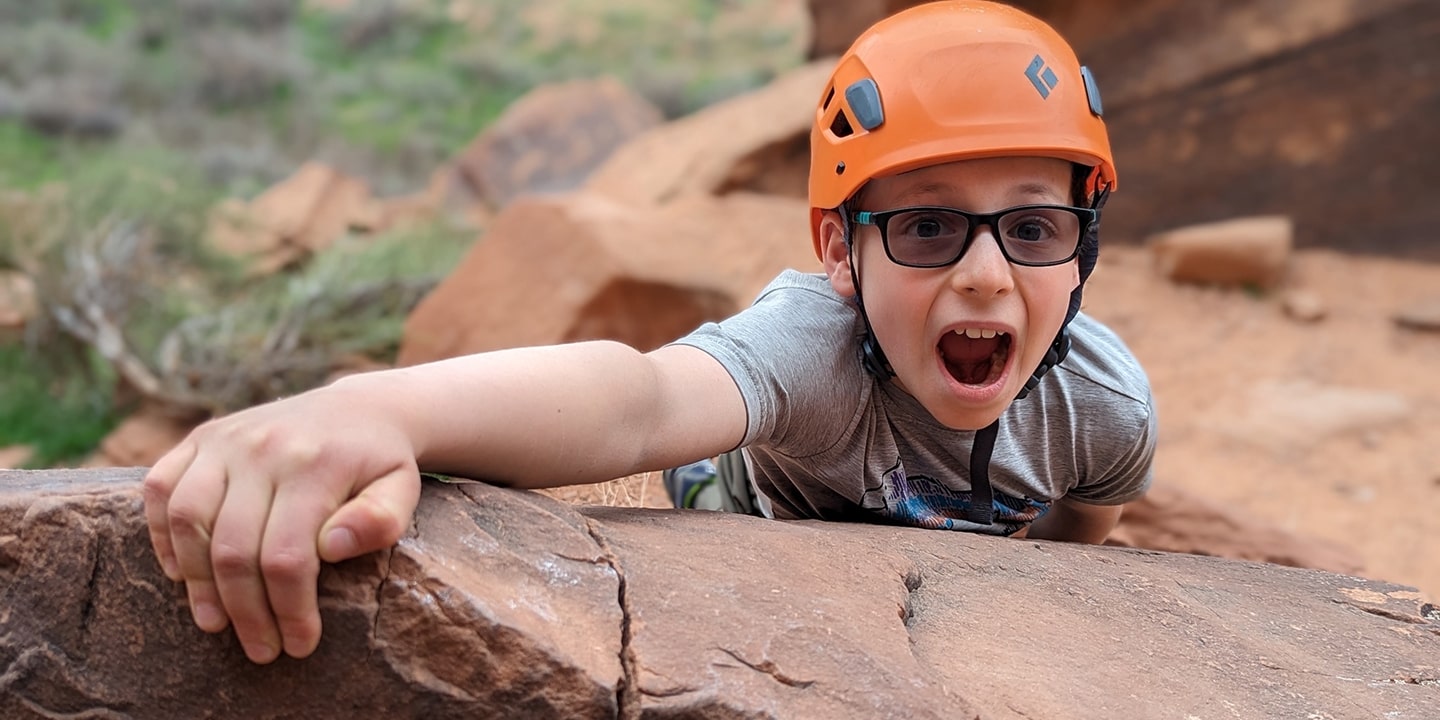 A kid in an orange helmet rock climbing with his mouth open